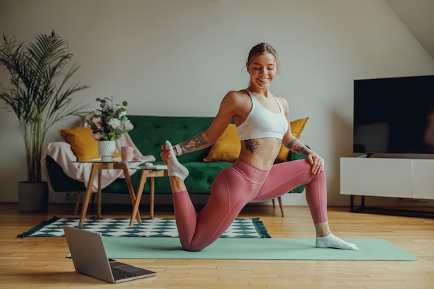 Una mujer sonriente está estirando las piernas en una alfombra de yoga frente a una computadora portátil Concepto de entrenamiento en casa