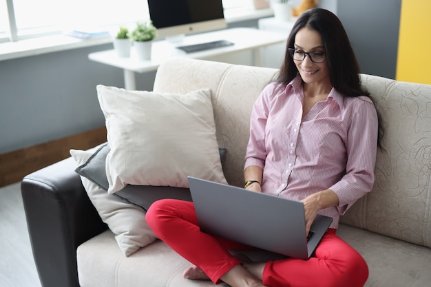 Mujer sonriente está sentada en el sofá y trabajando en la computadora portátil