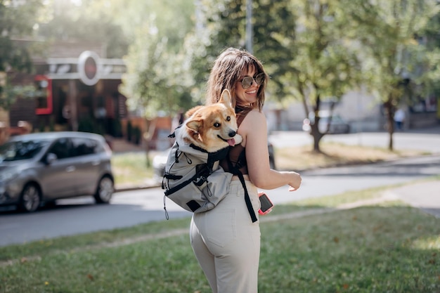 Mujer sonriente feliz caminando al aire libre en el parque de la ciudad con el perro Welsh Corgi Pembroke en una mochila especial