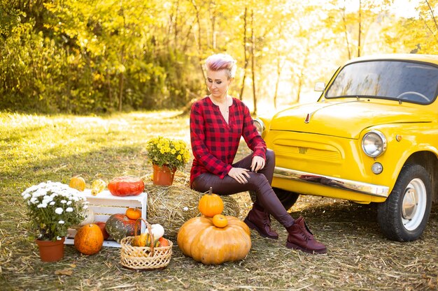 Mujer sonriente feliz cerca del coche amarillo con flores y calabazas en el fondo del parque de otoño con árboles dorados