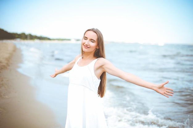 Mujer sonriente feliz en felicidad de felicidad libre en la playa del océano de pie con las manos abiertas. Retrato de una modelo femenina multicultural con vestido blanco de verano disfrutando de la naturaleza durante las vacaciones de viaje