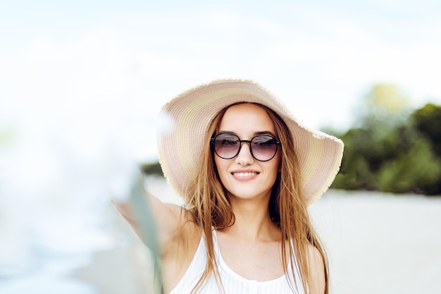 Mujer sonriente feliz en la felicidad de la felicidad libre en la playa del océano de pie con sombrero, gafas de sol y flores blancas. Retrato de una modelo femenina multicultural con vestido blanco de verano disfrutando de la naturaleza durante el viaje