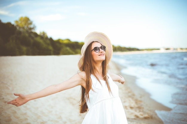 Mujer sonriente feliz en la felicidad de la felicidad libre en la playa del océano de pie con un sombrero, gafas de sol y las manos abiertas. Retrato de una modelo femenina multicultural con vestido blanco de verano disfrutando de la naturaleza durante el viaje