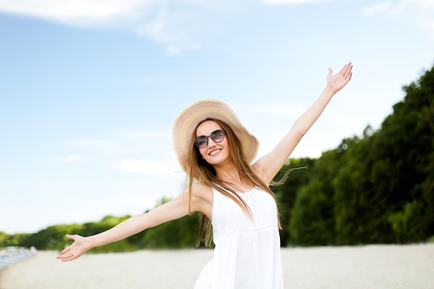 Mujer sonriente feliz en la felicidad de la felicidad libre en la playa del océano de pie con un sombrero, gafas de sol y las manos abiertas. Retrato de una modelo femenina multicultural con vestido blanco de verano disfrutando de la naturaleza durante el viaje
