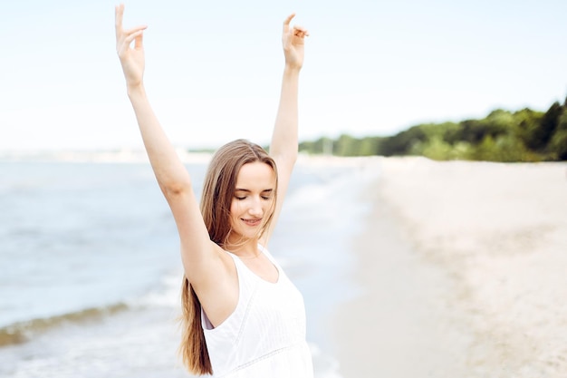 Mujer sonriente feliz en felicidad libre felicidad en la playa del océano de pie con las manos levantadas Retrato de una modelo femenina multicultural en vestido blanco de verano disfrutando de la naturaleza durante las vacaciones de viaje o