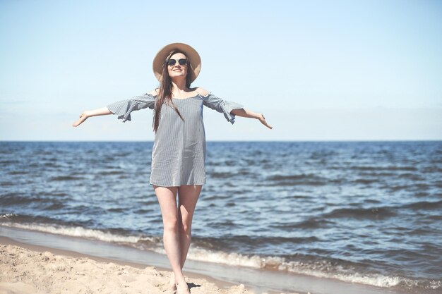 Mujer sonriente feliz en felicidad libre en la playa del océano de pie con las manos abiertas. Modelo de mujer morena con gafas de sol y sombrero disfrutando de la naturaleza durante las vacaciones de viaje al aire libre, plano medio.