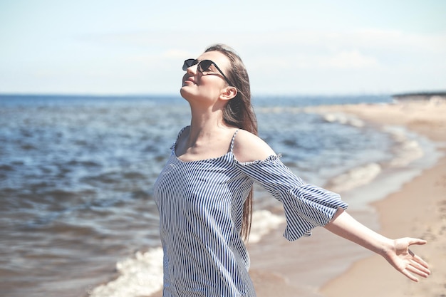 Mujer sonriente feliz en felicidad libre en la playa del océano de pie con las manos abiertas. Retrato de una modelo femenina morena vestida de verano disfrutando de la naturaleza durante las vacaciones de viaje al aire libre.