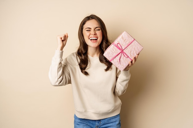 Mujer sonriente feliz sosteniendo un regalo y triunfando, levantando la mano alegremente, recibe un regalo de vacaciones, de pie sobre un fondo beige.