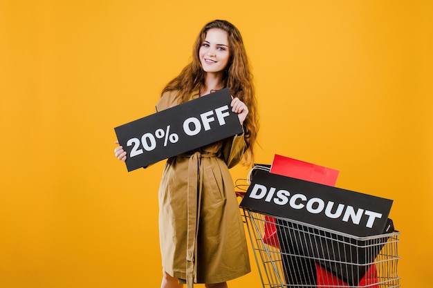 Mujer sonriente en gabardina con signo de descuento del 20% y coloridas bolsas de compras en carro aislado sobre amarillo