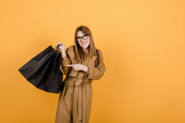 Mujer sonriente en gafas y gabardina con bolsas negras aisladas sobre amarillo