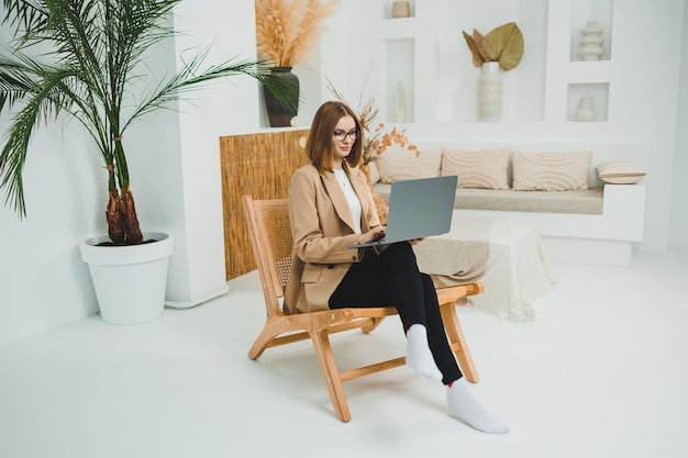 Mujer sonriente con gafas sentada en el sofá en casa relajada usando una computadora portátil trabajando remotamente en línea con camiseta blanca y vaqueros