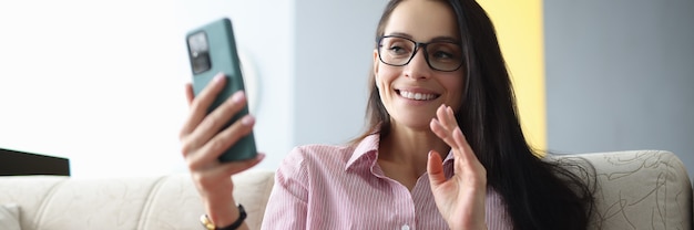 Mujer sonriente con gafas se sienta en el sofá, sostiene el teléfono inteligente y saluda a la pantalla.