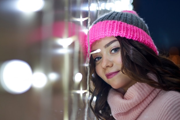 Mujer sonriente con guirnaldas y luces navideñas en Navidad festiva o año nuevo justo. Señora con suéter y mitones de punto de invierno clásico y elegante