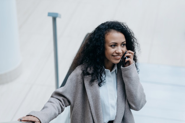 Mujer sonriente hablando por un teléfono inteligente de pie en una escalera mecánica en un centro comercial