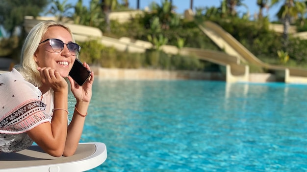 Mujer sonriente hablando por teléfono en una tumbona cerca de la piscina. Copia espacio