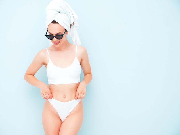 Mujer sonriente hermosa joven en ropa interior blanca. Sexy modelo despreocupado en ropa interior y toalla en la cabeza posando junto a la pared azul claro en el estudio. Mujer positiva y feliz disfrutando de la mañana con gafas de sol