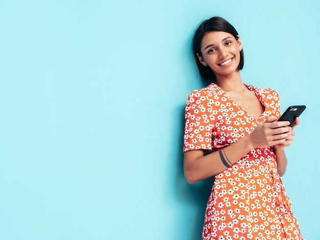 Mujer sonriente hermosa joven en vestido rojo de verano de moda Mujer despreocupada sexy posando cerca de la pared azul en el estudio Modelo mirando la pantalla del teléfono celular Sosteniendo el teléfono inteligente y usando aplicaciones