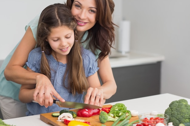 Mujer sonriente con hija cortar verduras en la cocina