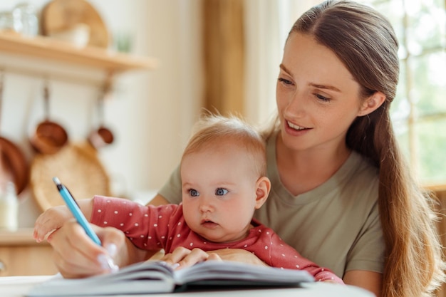 mujer sonriente independiente sosteniendo un bebé lindo escribiendo notas mientras trabaja desde casa concepto de paternidad