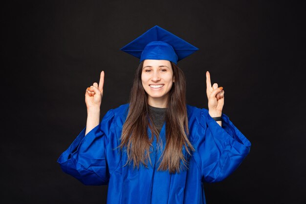 Mujer sonriente joven alegre vistiendo soltero y gorro de graduación y apuntando hacia arriba