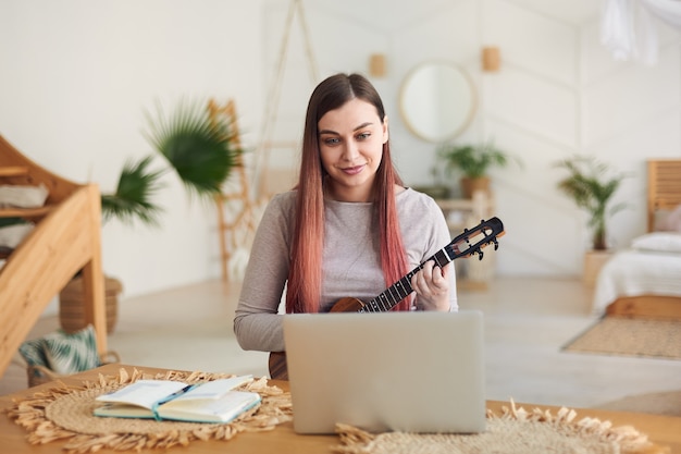 Mujer sonriente joven aprendiendo a tocar el ukelele de forma remota. El profesor da una lección de ukelele en línea.