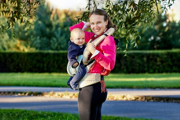 Foto mujer sonriente joven con bebé en cabestrillo para bebé camina en el parque durante la licencia de maternidad.