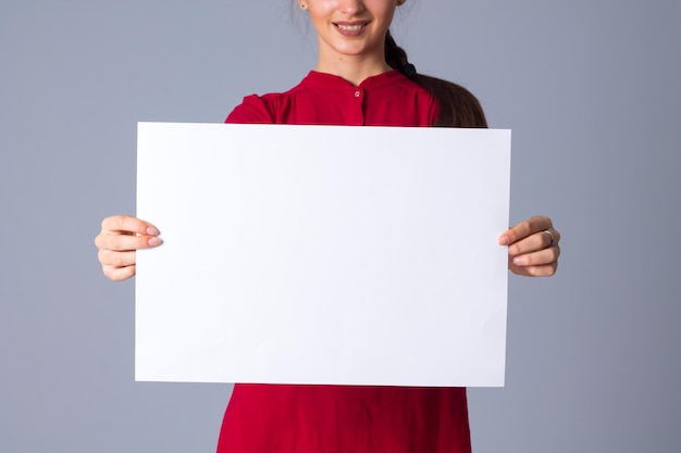 Mujer sonriente joven en blusa roja con trenza que muestra una hoja blanca de papel en el estudio