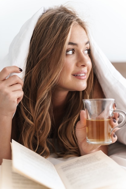 Foto mujer sonriente joven con cabello rubio leyendo un libro y bebiendo té mientras está acostado debajo de una manta después de dormir