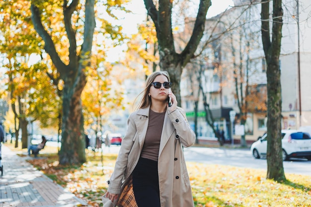 Mujer sonriente joven caminando en el parque de otoño hablando por teléfono inteligente con gafas de sol de gabardina tendencia de estilo de moda de humor feliz