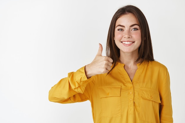 Mujer sonriente joven en camisa amarilla muestra los pulgares para arriba en aprobación, pared blanca