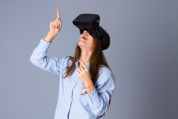 Mujer sonriente joven en camisa azul usando gafas VR y mirando hacia arriba sobre fondo gris en estudio