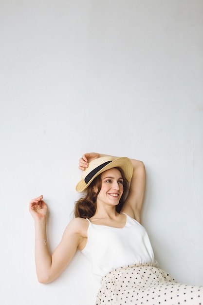 Mujer sonriente joven en camisa blanca y sombrero sentado sobre fondo blanco.