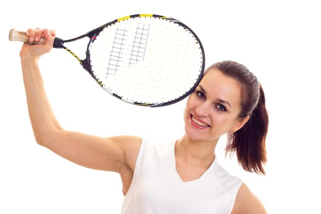 Mujer sonriente joven en camisa deportiva blanca con cola de caballo oscura con raqueta de tenis en estudio