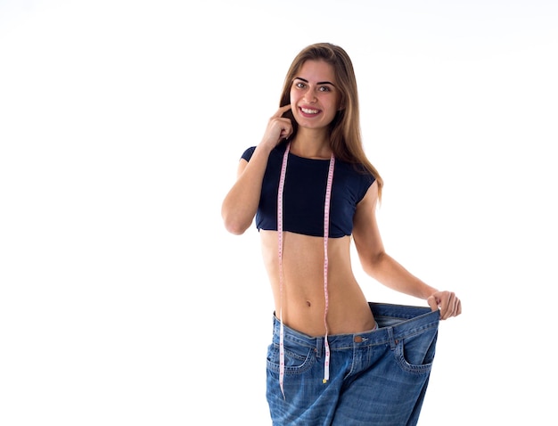 Mujer sonriente joven con centímetro alrededor de su cuello sosteniendo sus jeans de tamaño mucho más grande