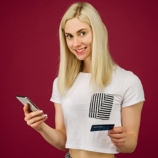 Mujer sonriente joven compra en línea. Tiene un teléfono inteligente y una tarjeta de crédito en la mano en ruby