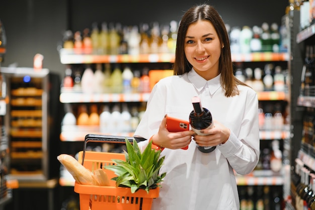 Mujer sonriente joven comprando en un supermercado mirando la pantalla del teléfono su lista de compras (lista de compras) - consumismo y concepto de elección