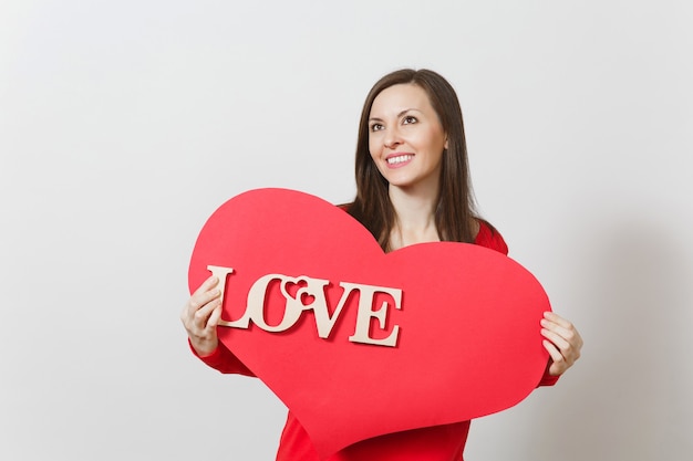 Mujer sonriente joven eficaz en ropa casual roja que sostiene el corazón rojo grande, amor de la palabra de madera en el fondo blanco. Copie el espacio para publicidad. Concepto de día de San Valentín o día internacional de la mujer.