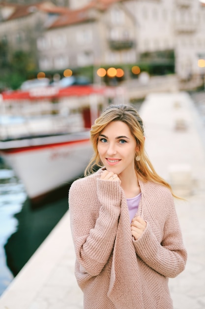 Mujer sonriente joven envuelta en una chaqueta abrigada mientras está de pie en el muelle con el telón de fondo de