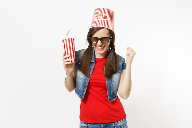 Mujer sonriente joven en gafas 3d con balde para palomitas de maíz en la cabeza viendo una película sosteniendo un vaso de plástico de refresco o cola haciendo el cartel ganador diciendo Sí aislado sobre fondo blanco. Emociones en el cine.