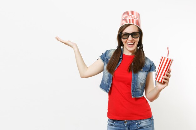 Mujer sonriente joven en gafas 3d con cubo para palomitas de maíz en la cabeza viendo una película sosteniendo una taza de refresco o cola apuntando con la mano a un lado en el espacio de la copia aislado sobre fondo blanco. Emociones en el cine.