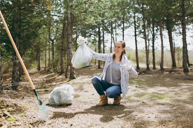 Mujer sonriente joven limpiando basura sosteniendo bolsas de basura mostrando el pulgar hacia arriba en el parque o bosque