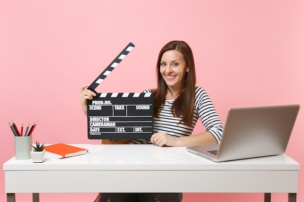 Mujer sonriente joven mantenga claqueta de cine negro clásico trabajando en el proyecto mientras se sienta en la oficina con la computadora portátil