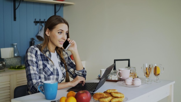 Mujer sonriente joven navegando en las redes sociales usando una computadora portátil y hablando por teléfono