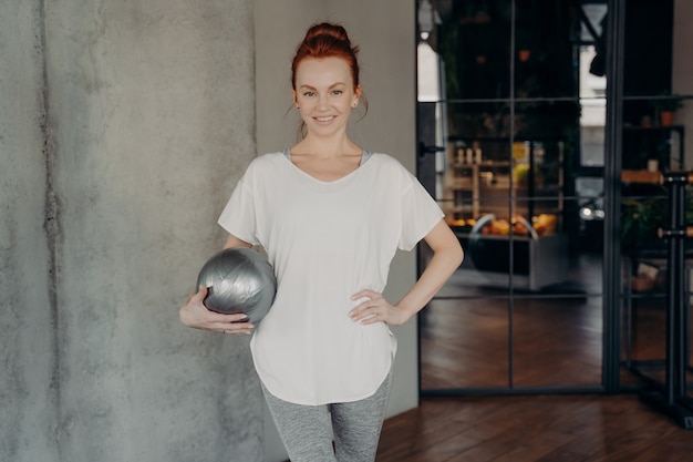 Mujer sonriente joven pelirroja de pie con fitball en una mano viste camiseta deportiva blanca y leggings grises listos para el entrenamiento de barra en el gimnasio. Estilo de vida saludable y concepto interior de pilates.