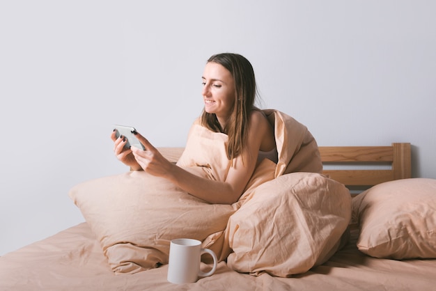 Mujer sonriente joven que disfruta usando el teléfono inteligente mientras está sentado en la cama cómoda por la mañana