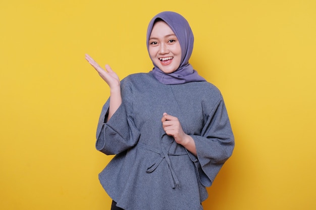 Mujer sonriente joven que lleva el hijab muestra algo y sostiene algo con las palmas aisladas sobre fondo amarillo claro de la bandera