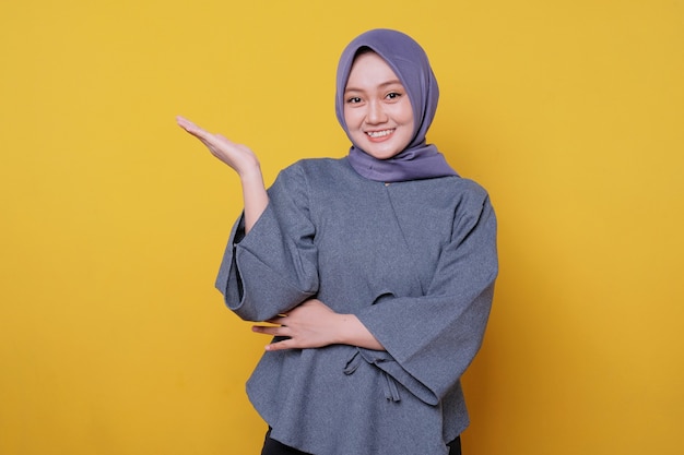 Mujer sonriente joven que lleva el hijab muestra algo y sostiene algo con las palmas aisladas sobre fondo amarillo claro de la bandera