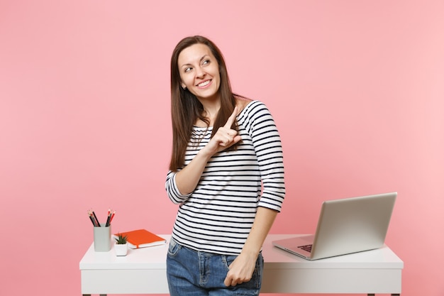 Mujer sonriente joven que señala el dedo índice a un lado del trabajo y de pie cerca del escritorio blanco con computadora portátil pc