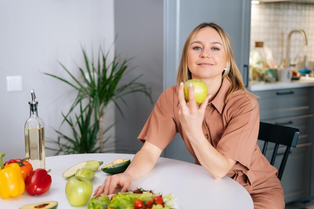 Mujer sonriente joven que sostiene la manzana en la cocina en la cocina con un interior moderno. Concepto de alimentación saludable.