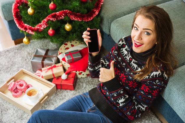 Mujer sonriente joven que sostiene el teléfono y que muestra el pulgar para arriba en Navidad